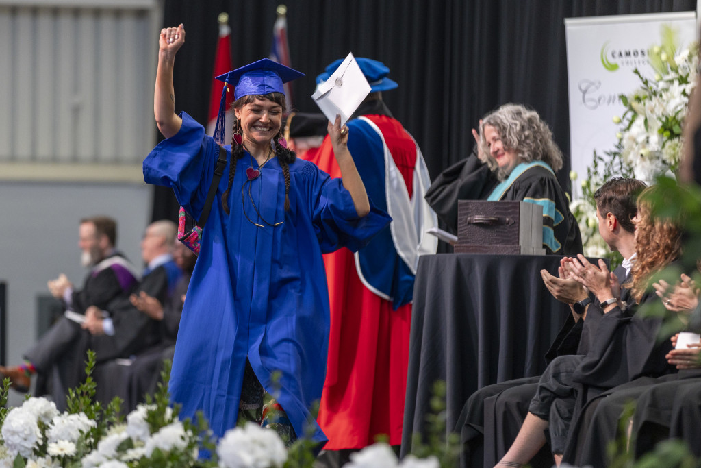 A woman walks across the convocation stage in grad regalia with her arms in the air