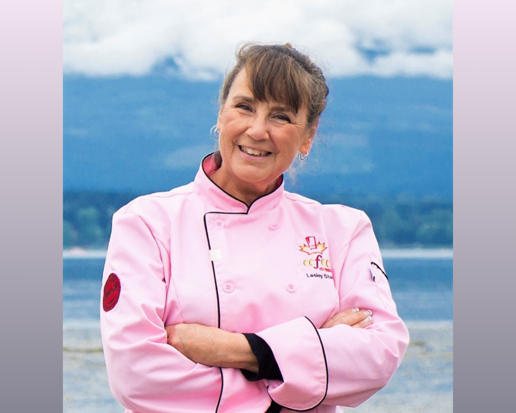 A woman in a pink chef's jacket stands in front of ocean and mountains.