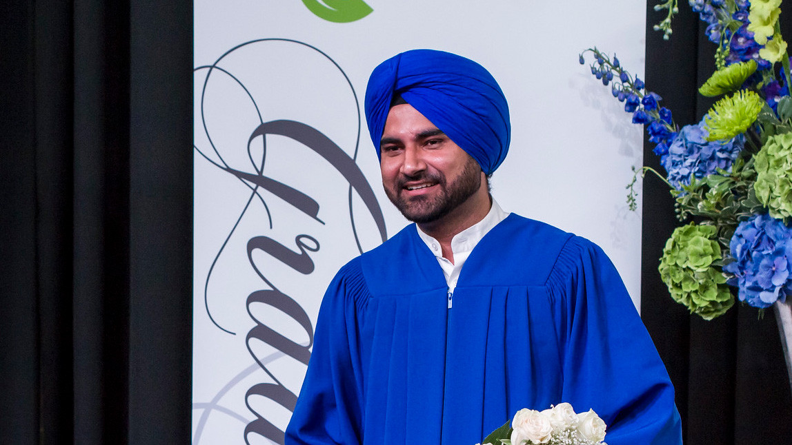 A man in a graduation robe wears a matching turban and smiles for the camera