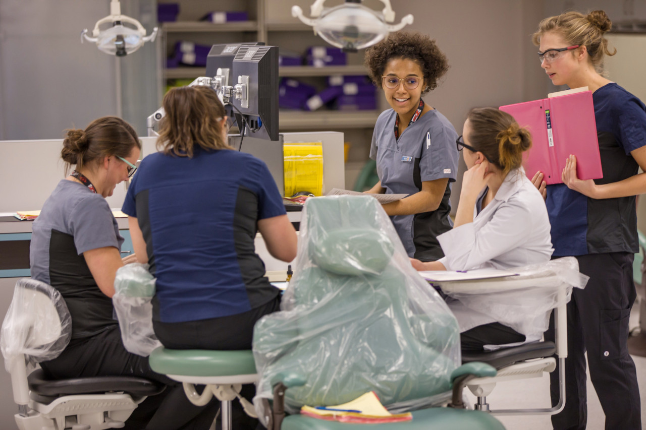 A group of 5 dental studnts working together in a practice lab. They are laughing and chatting as they work on an assignment.