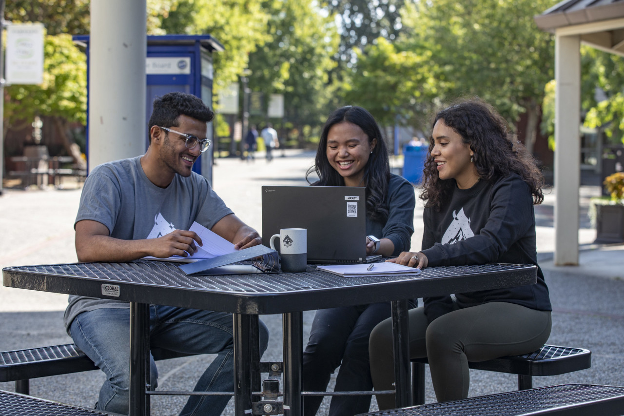 3 students outside at a table using a laptop for study