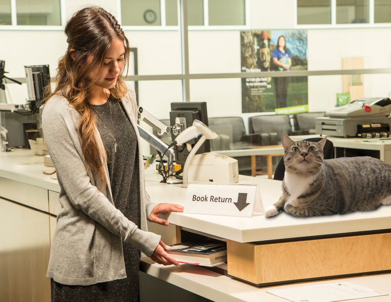 A young woman places a text book in the slot marked 'book return'. A photshopped cat is on top of the counter and appears to be looking at the camera.