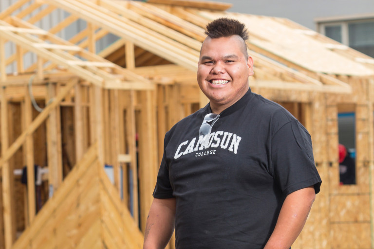 An Indigenous Building Trades student stands in front of a house that is only framed in