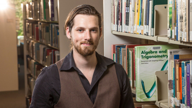 A young man standing in the Library looking at Algebra textbook